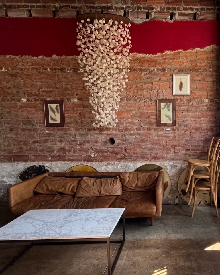 A draping piece of art hangs above a brick wall with three framed pictures on it and a leather couch below inside Red Tree Coffee and Art in Oakley