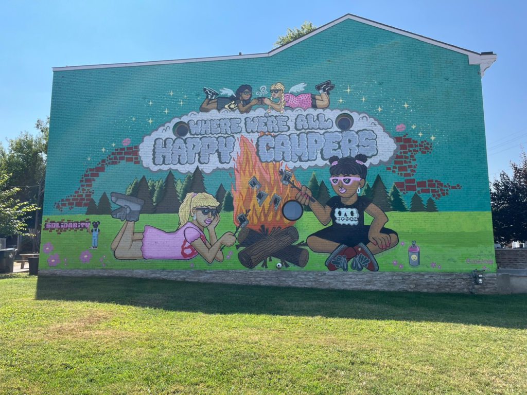 Mural of two girls at a camp fire with the words "Where we're all happy campers" on the outer wall of Mom 'N 'Em in Camp Washington
